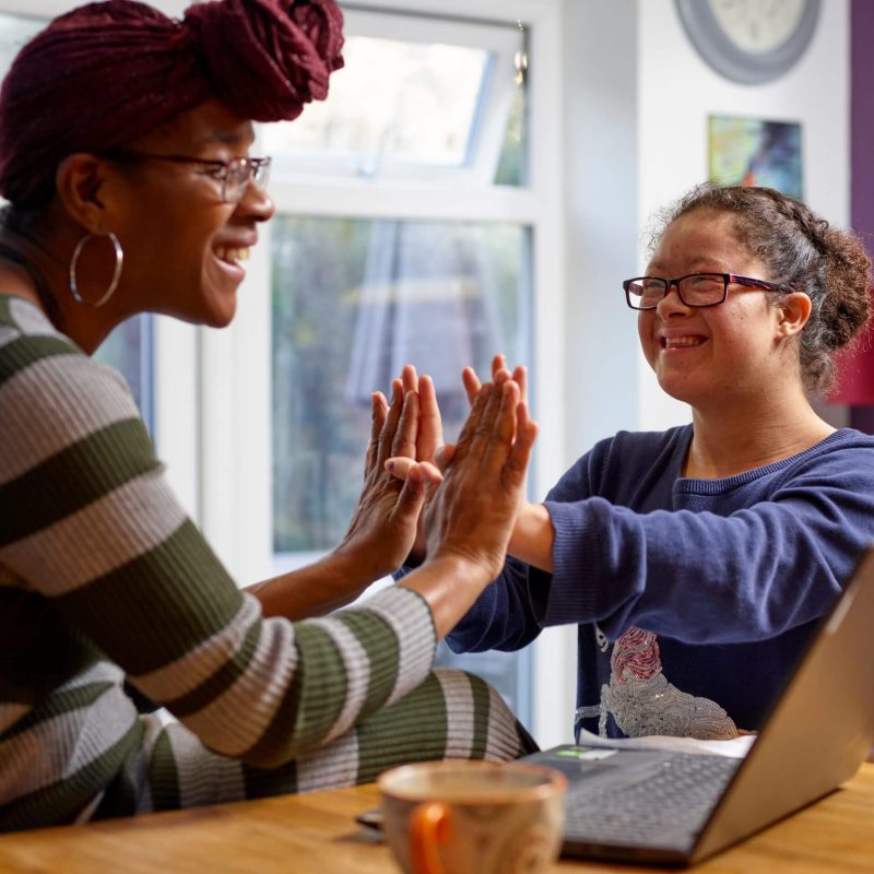 support-worker-and-young-girl-living-with-down-syndrome-playing-cheerfully-at-home-in-front-of-computer-clapping-hands-scaled-1